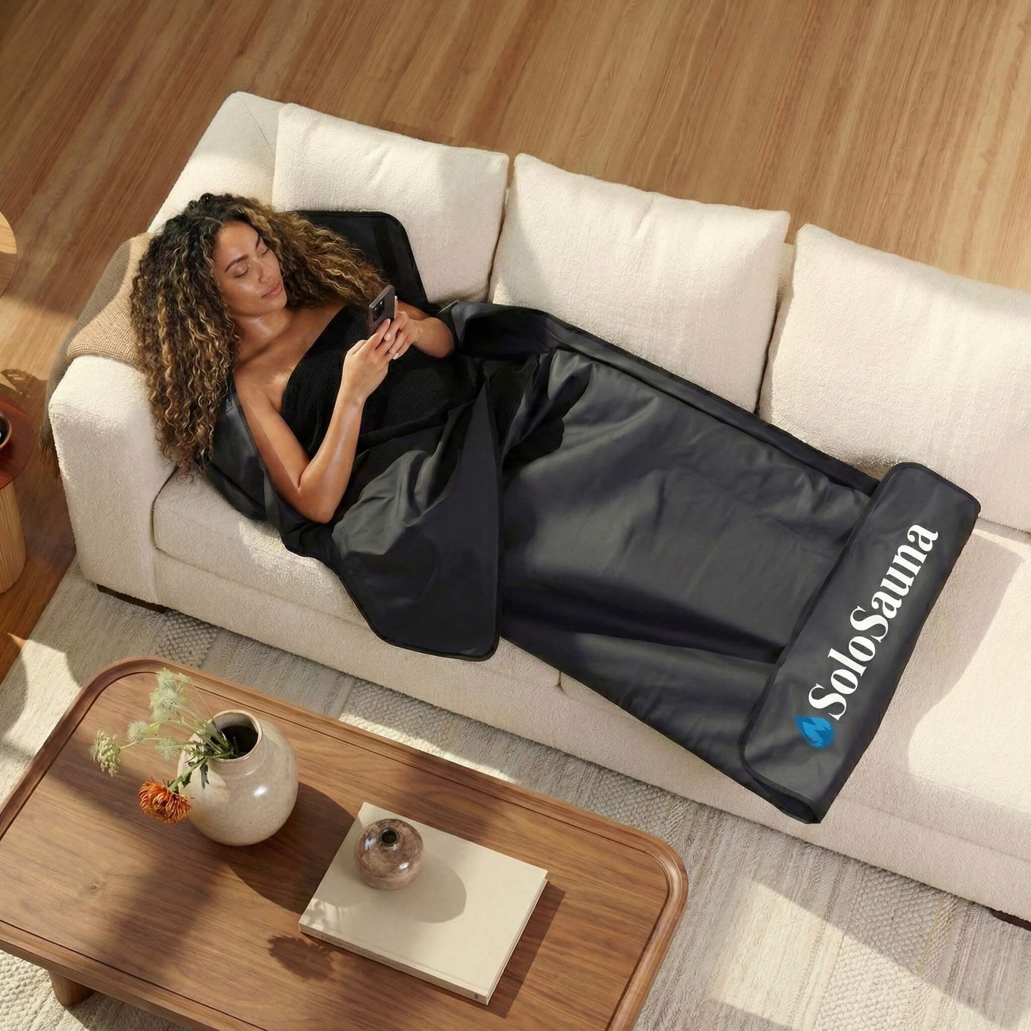 Woman lying on a couch with a black sauna blanket, using a phone, in a home setting.