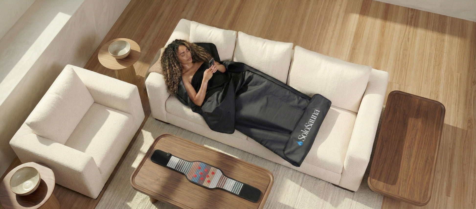 Woman lying on a white couch with a black blanket, surrounded by wooden coffee tables and a white chair.
