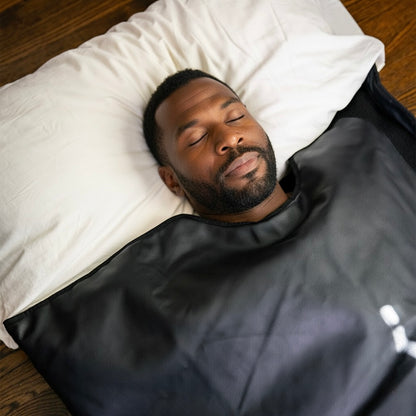 Man sleeping under a black sauna blanket on a wooden floor