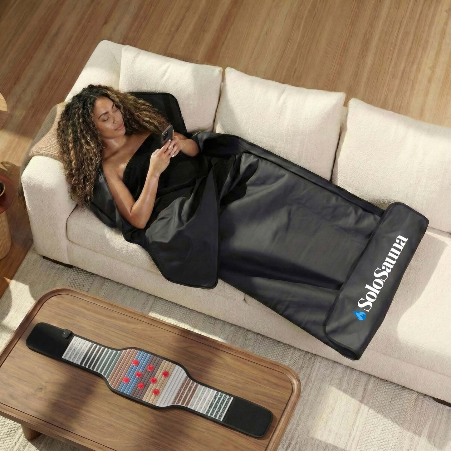 A woman is in a SoloSauna blanket on a couch with an infrared belt on the table beside her.